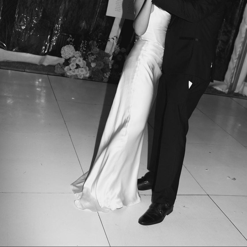 A black and white photograph of a bride in a sleeveless satin wedding gown and a groom in a dark suit dancing on a dance floor inside a decorated event tent with string lights.