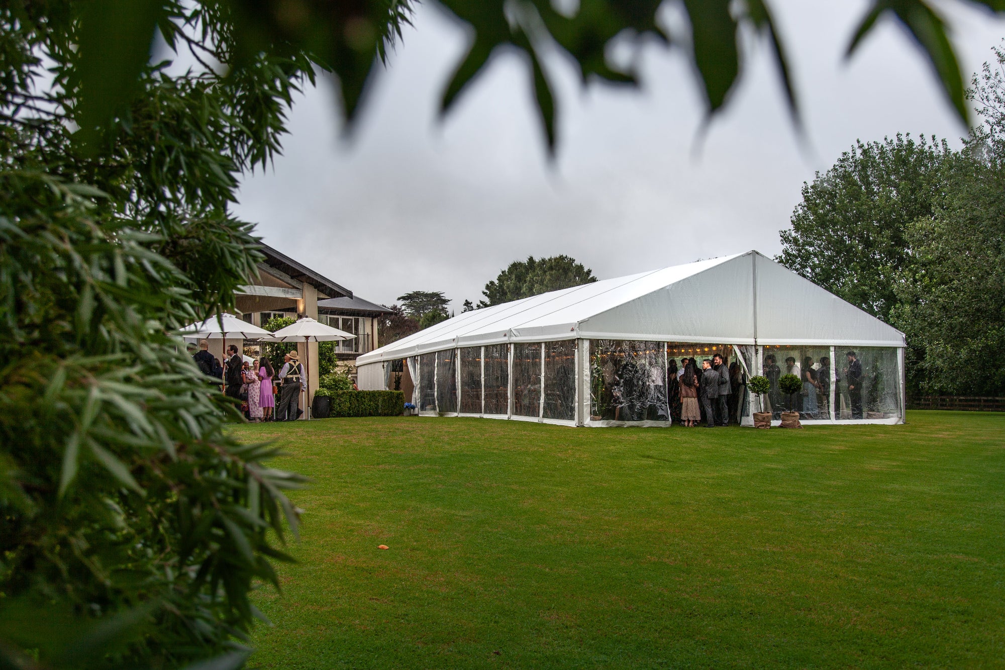 A large white event marquee tent with clear side walls is set up on a green lawn outdoors, with guests visible inside and trees and a building in the background under an overcast sky.